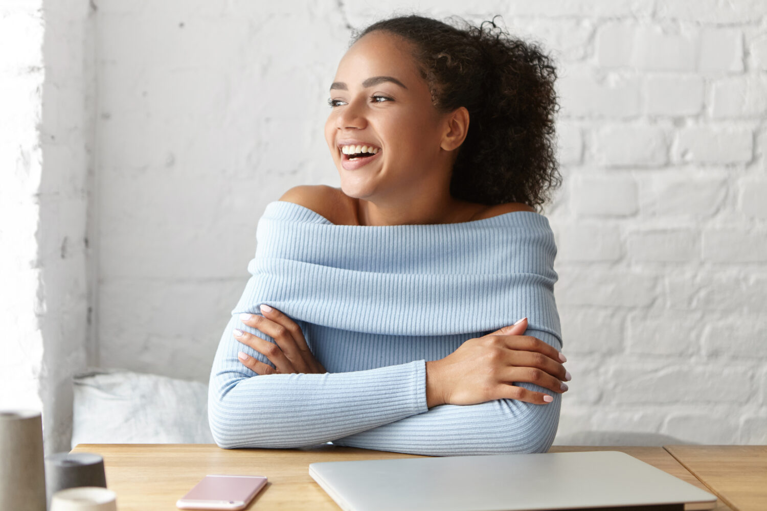Adorable dark-skinned young woman with Afro hairstyle keeping her hands crossed while sitting at wooden table surrounded with modern technologies, having pleased expression while looking aside